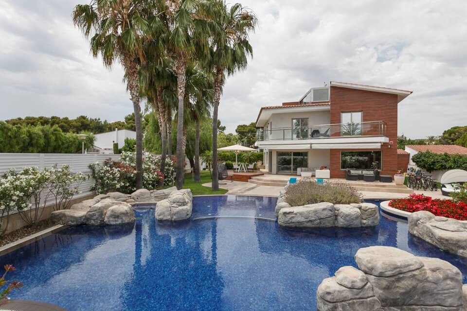Vistas de la casa y la piscina decorada con piedra artificial, con grupo de palmeras en el jardín y bosque de pinos al fondo.